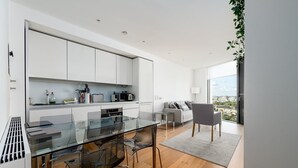 Une cuisine moderne avec des meubles blancs, une table à manger en verre et une vue sur la ville grâce à de grandes fenêtres.