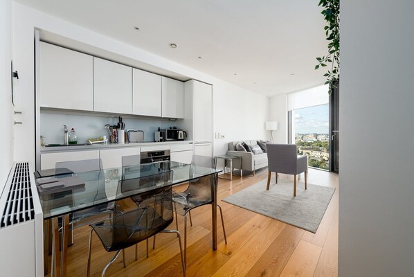 Une cuisine moderne avec des meubles blancs, une table à manger en verre et une vue sur la ville grâce à de grandes fenêtres.