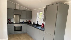 A modern kitchen with grey cabinets, a built-in oven, a coffee machine, and a window with blinds. - Tranquil Lodge Retreat in Wells (Wells)