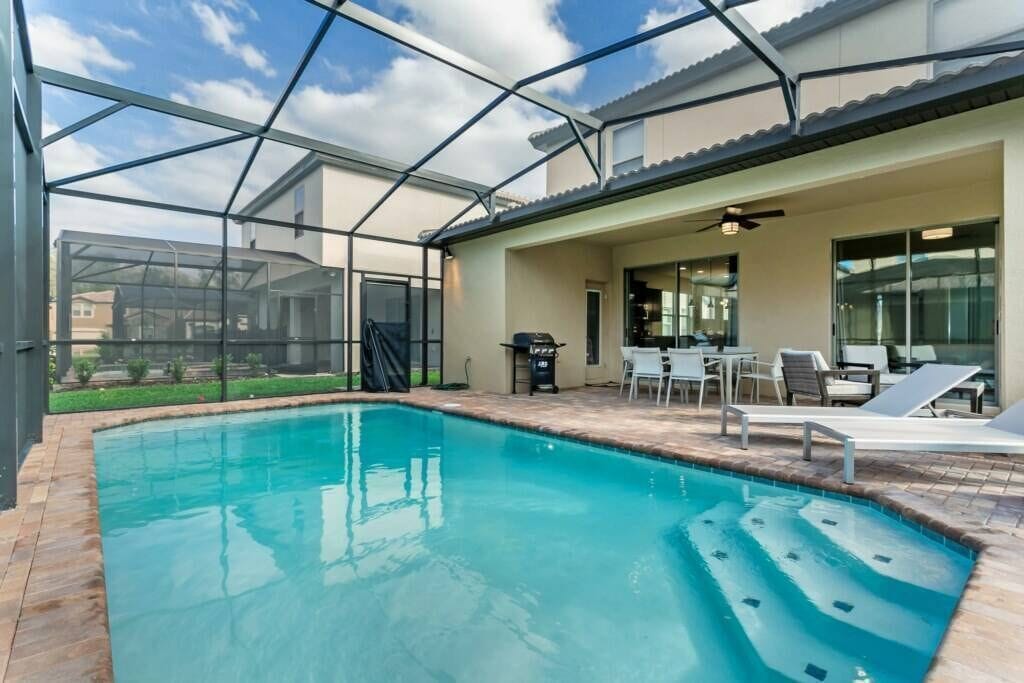 A swimming pool with a glass enclosure, a patio area with chairs and a table, and a view of a residential neighborhood.