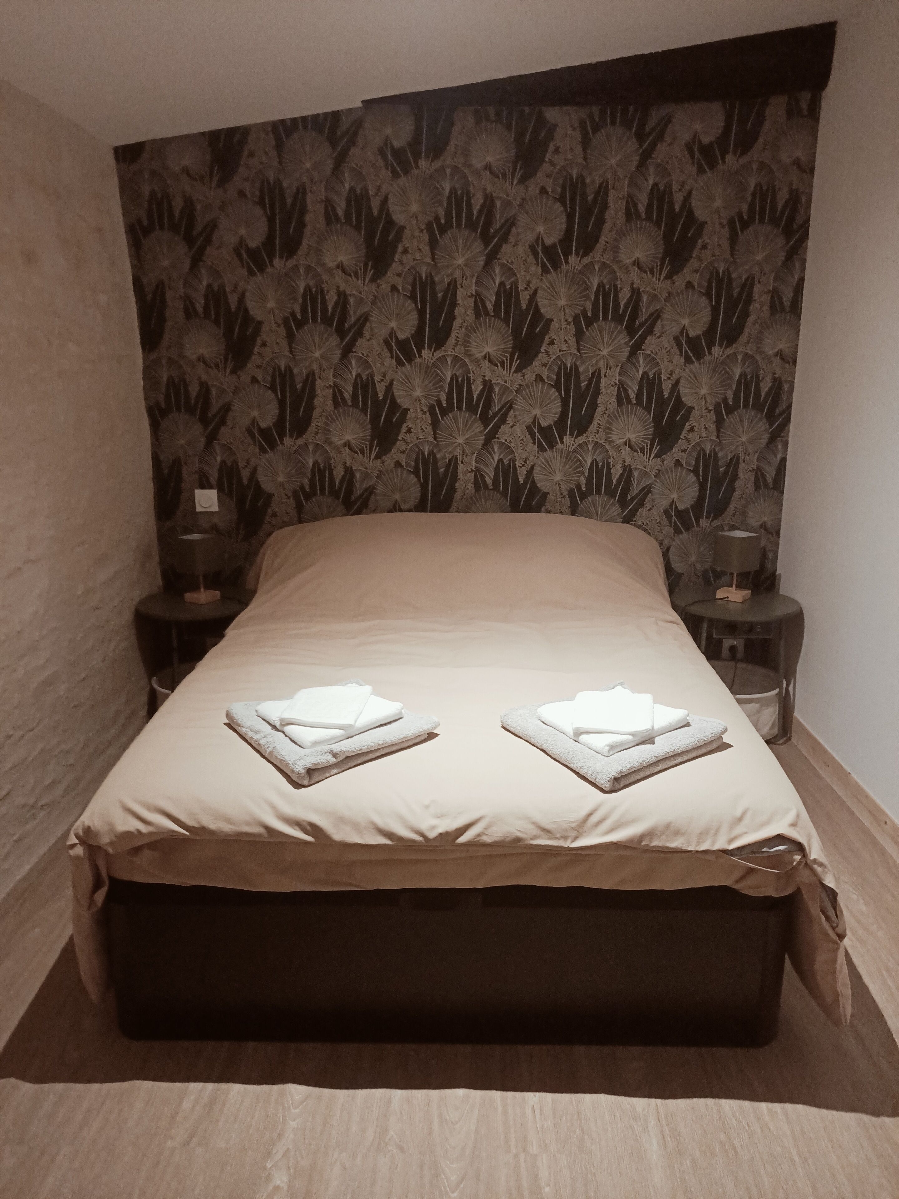 A neatly made bed with a beige cover and two folded towels on the footboard. The room has a dark floral wallpaper and two bedside tables with lamps.