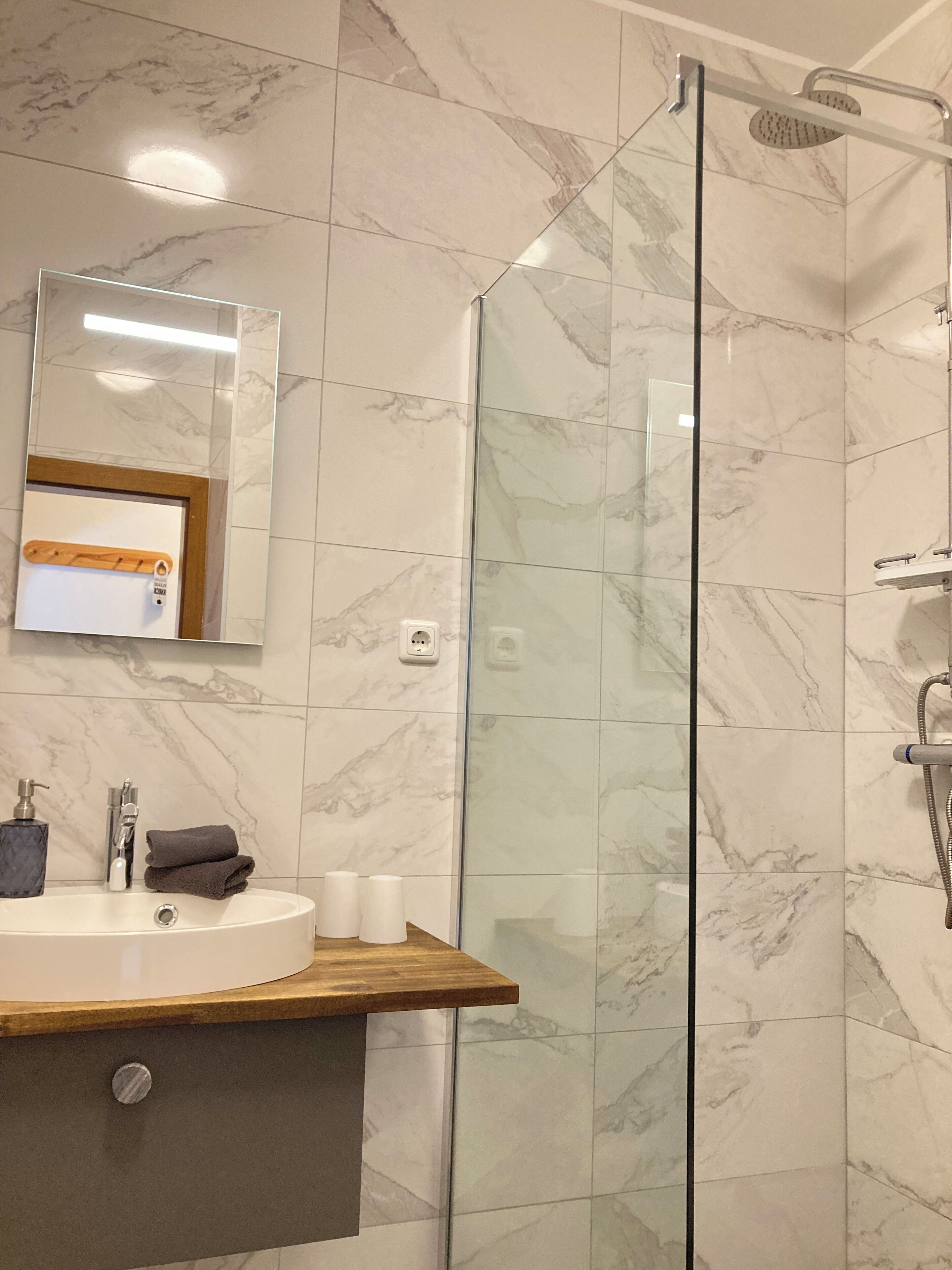 Un bagno moderno con cabina doccia in vetro, piastrelle in marmo, mobile bagno in legno, lavandino e specchio.
