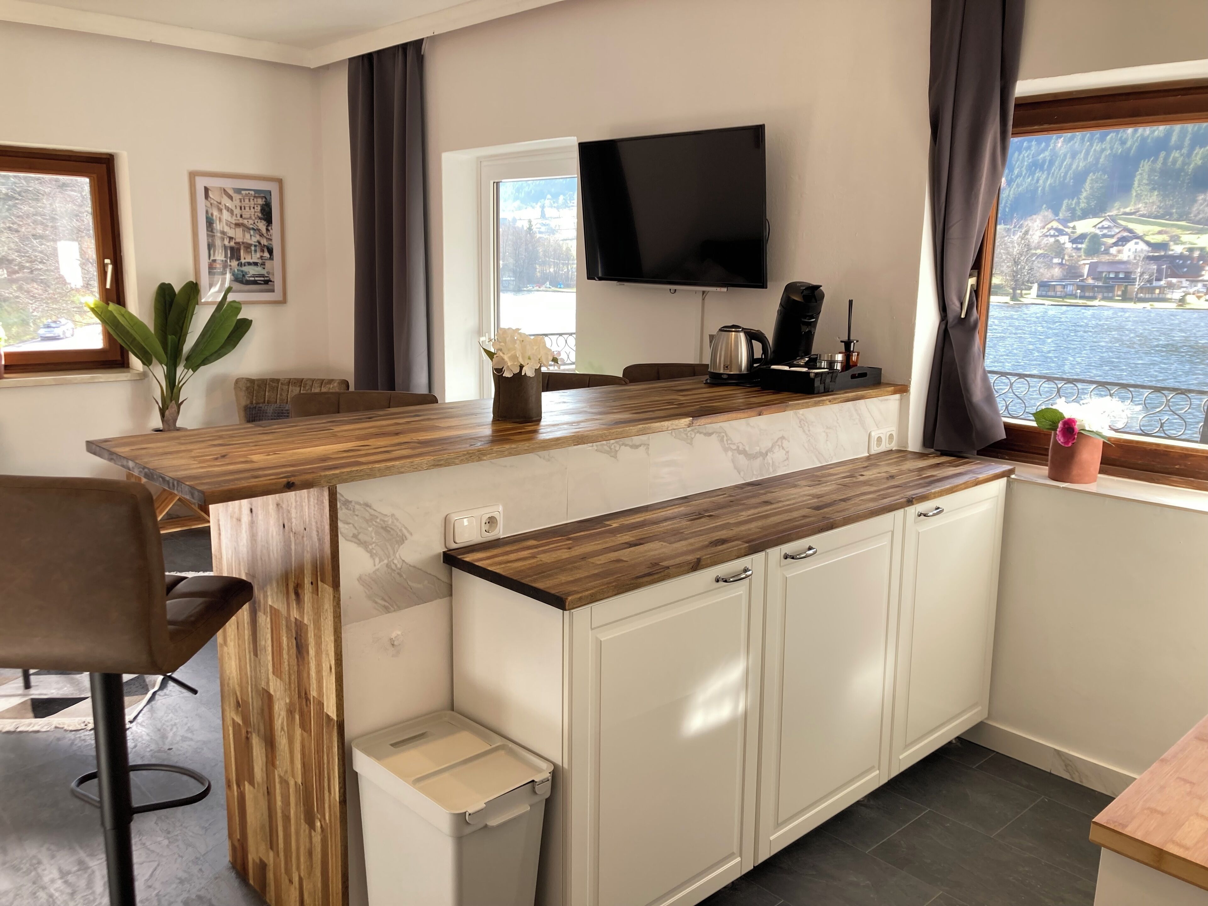 A modern kitchen with a wooden countertop, white cabinets, a built-in TV, and a view of a lake.