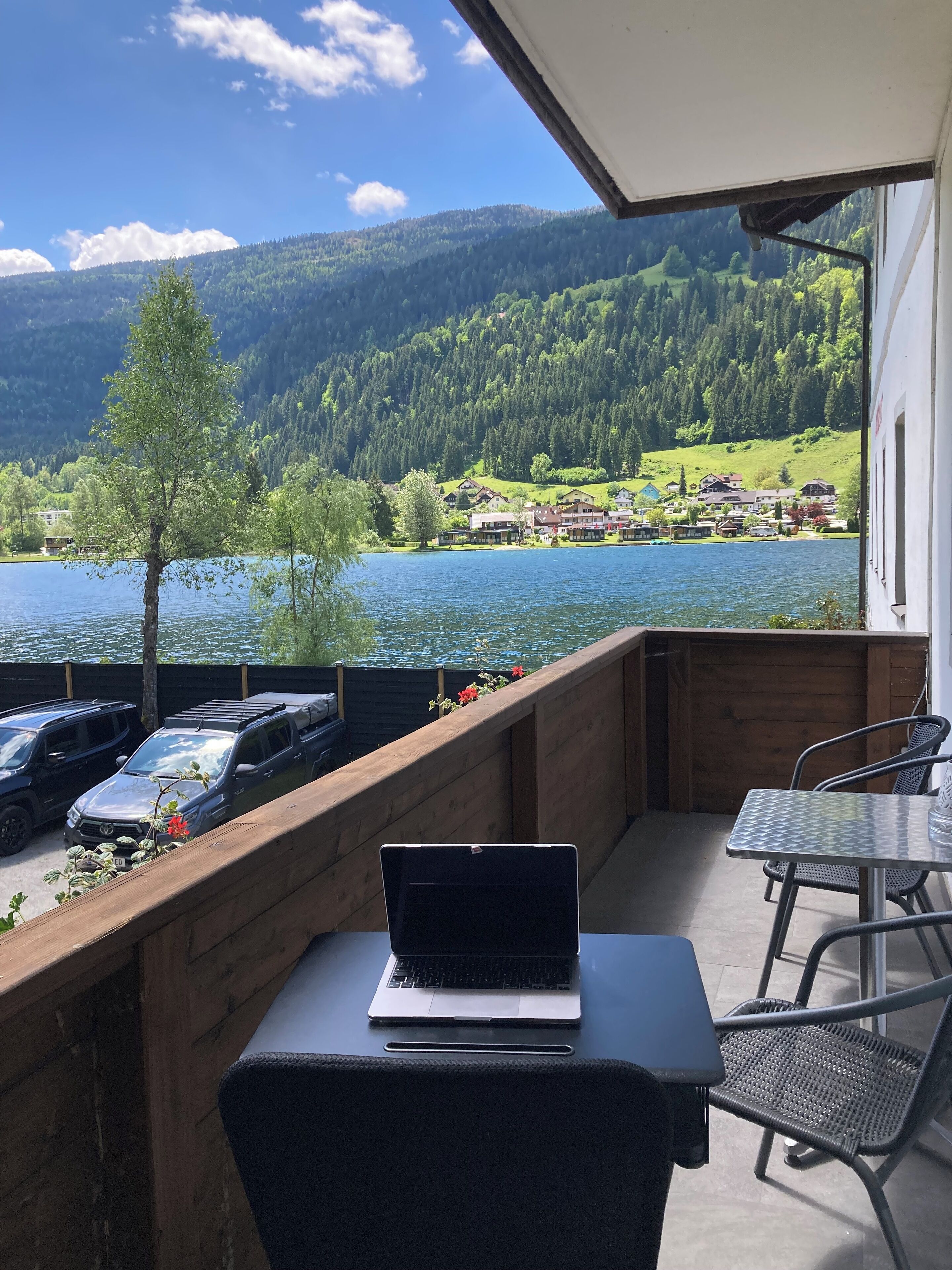 En balkon med en bærbar computer, to stole og et bord med udsigt over en sø og skovklædte bakker.