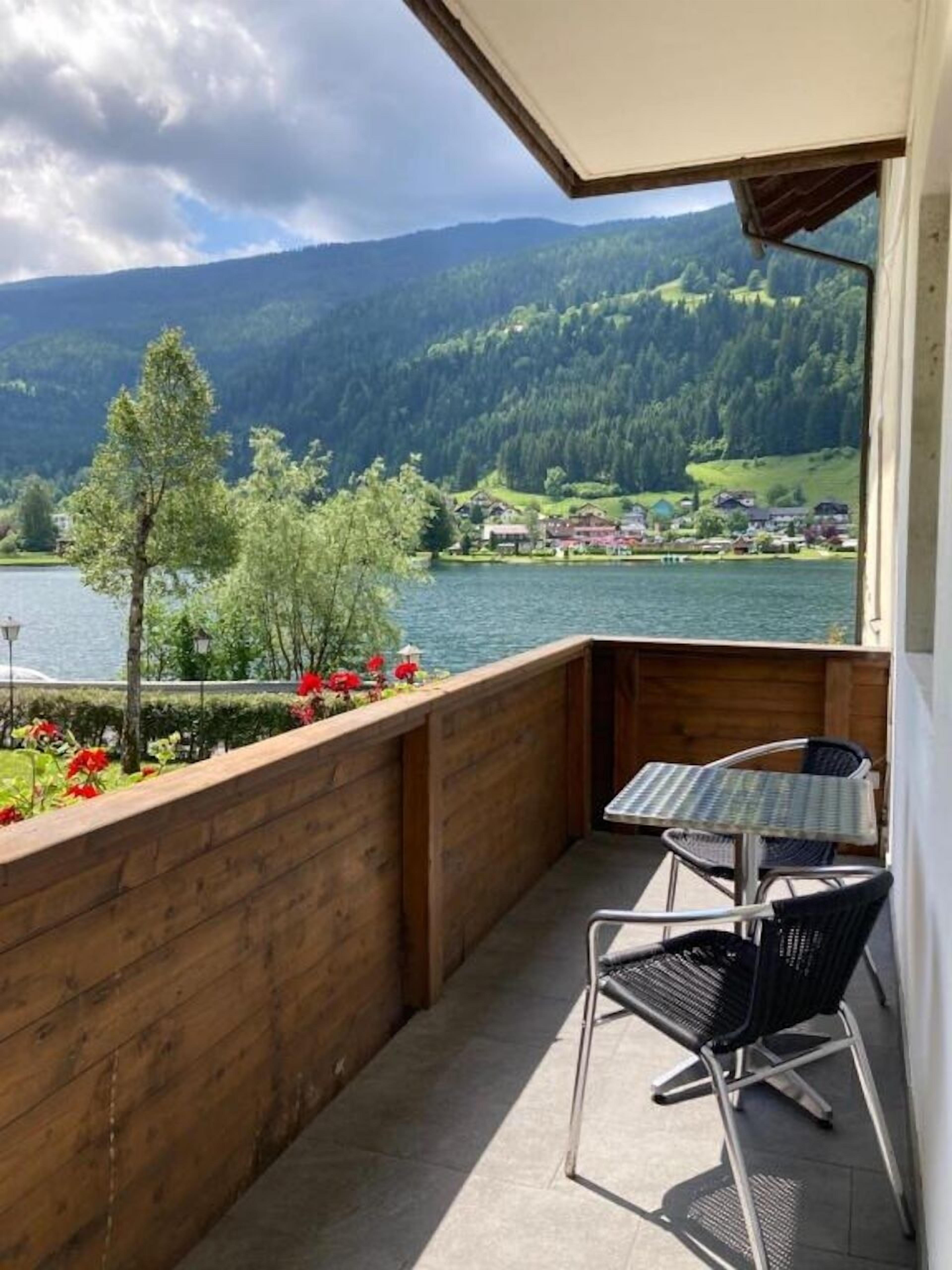 A balcony with a view of a lake and mountains, wooden railing, and a table with chairs.