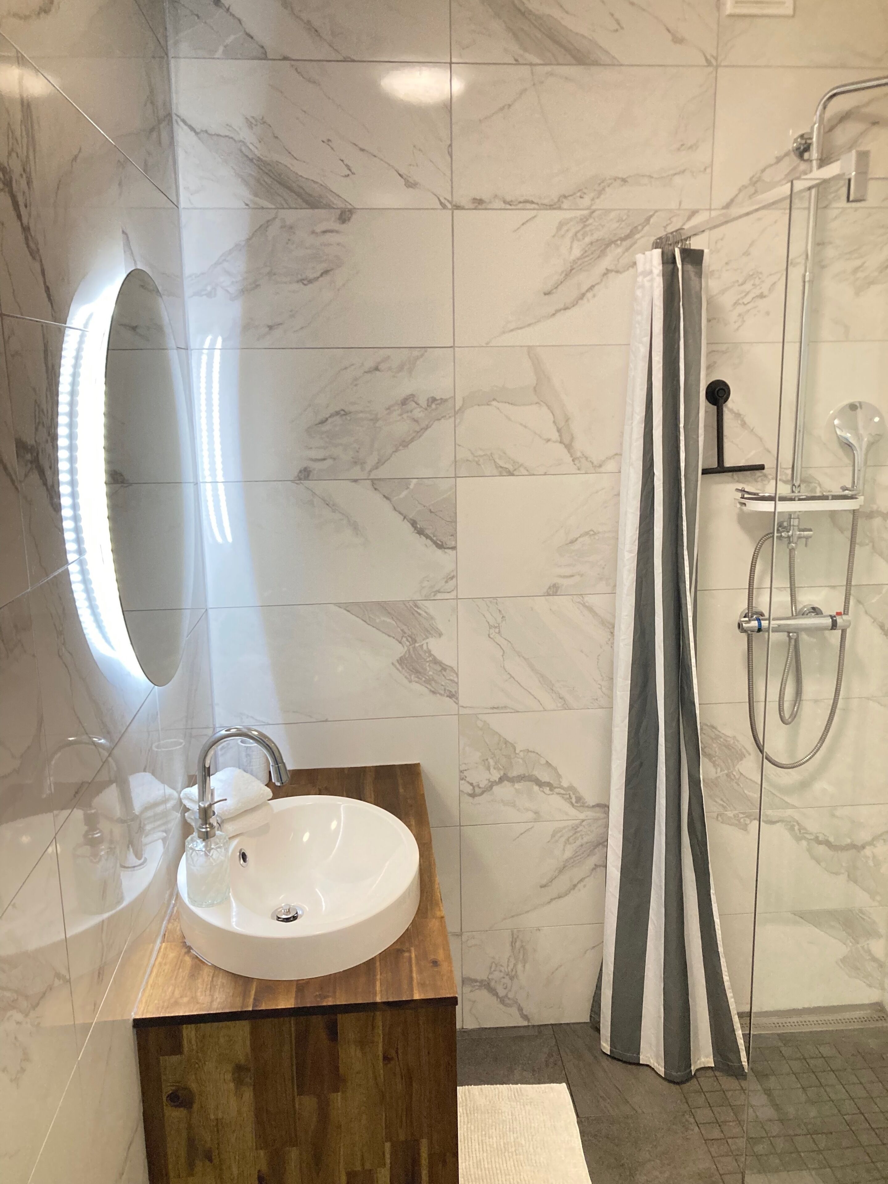 Un baño con piso de mármol, tocador de madera con lavamanos, espejo y zona de ducha con cortina a rayas.