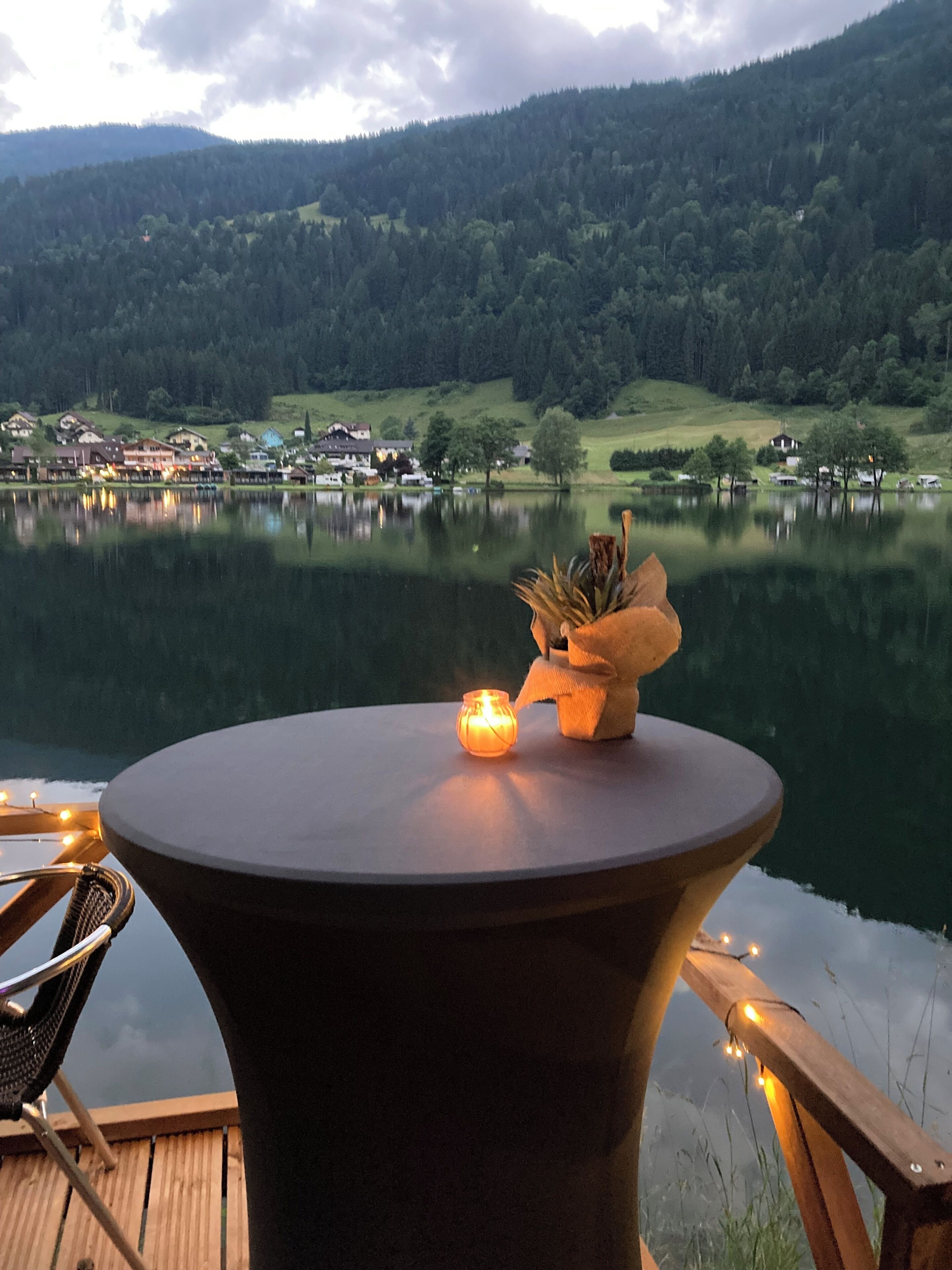 Ein Tisch am See mit Kerze und dekorativer Vase, mit Blick auf einen ruhigen See und einen bewaldeten Hügel mit Gebäuden im Hintergrund.