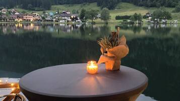 Ein Tisch am See mit Kerze und dekorativer Vase, mit Blick auf einen ruhigen See und einen bewaldeten Hügel mit Gebäuden im Hintergrund.