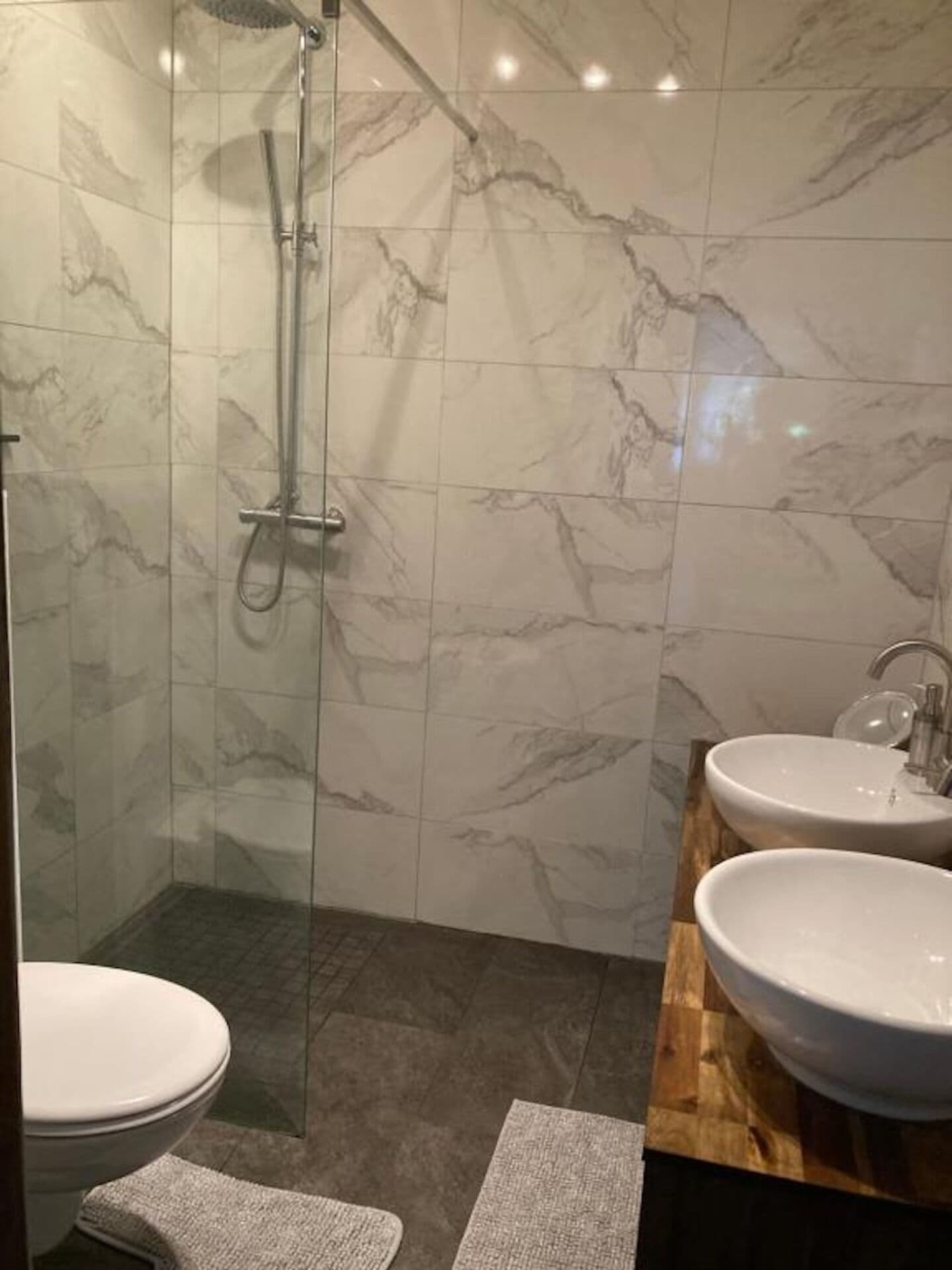Un baño con paredes de mármol, cabina de ducha con mampara de vidrio, dos lavabos y un inodoro.