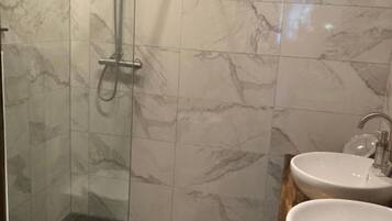 Un baño con paredes de mármol, cabina de ducha con mampara de vidrio, dos lavabos y un inodoro.