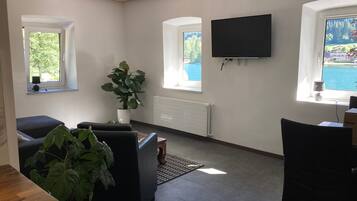 Ein modernes Wohnzimmer mit einem an der Wand befestigten Flachbildfernseher, einer großen Kühlpflanze und Seeblick durch die Fenster.