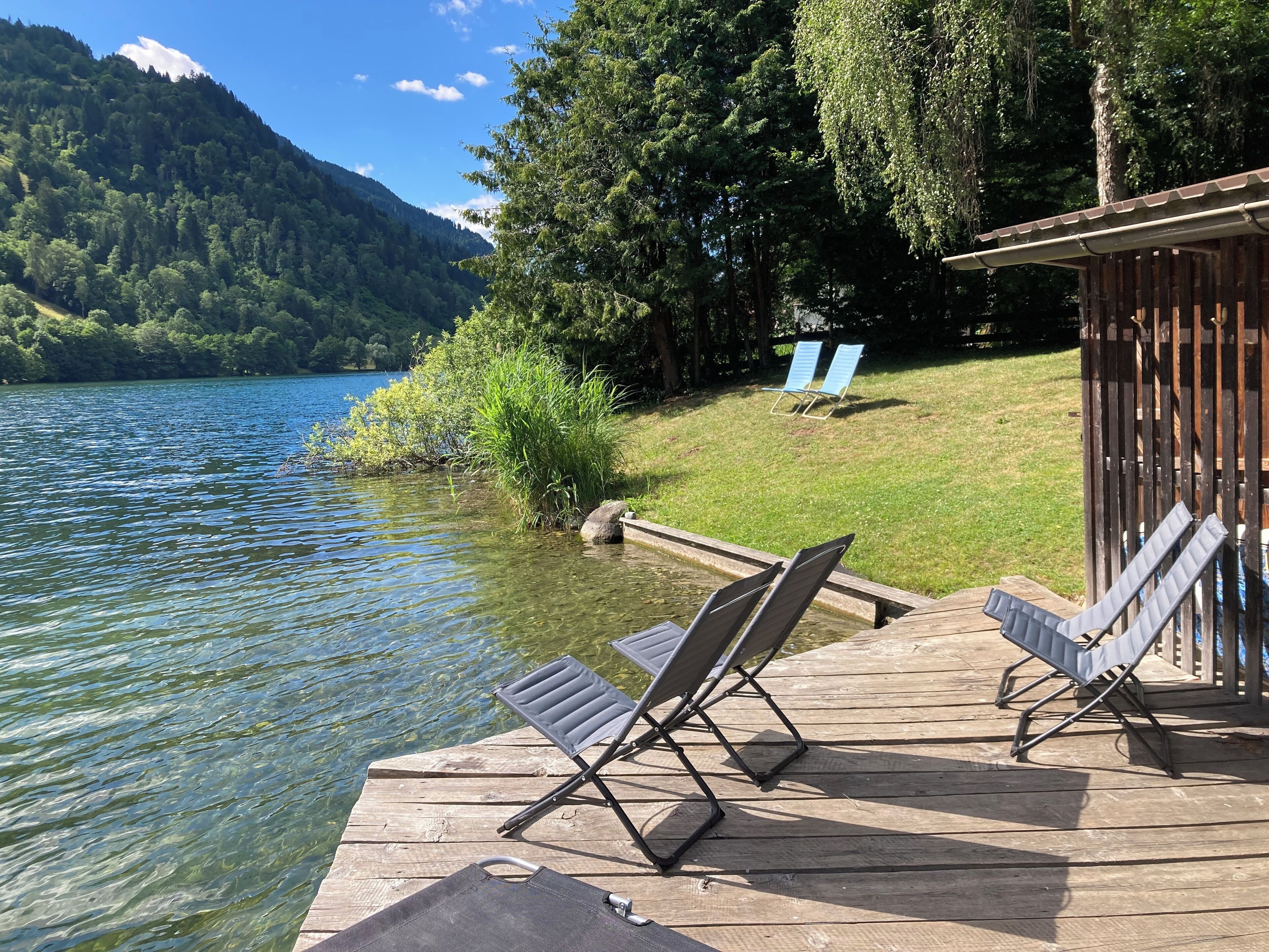 Eine Holzterrasse mit zwei Liegestühlen, die auf einen See ausgerichtet sind, umgeben von viel Grün und einem Berg im Hintergrund.