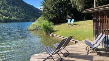Eine Holzterrasse mit zwei Liegestühlen, die auf einen See ausgerichtet sind, umgeben von viel Grün und einem Berg im Hintergrund.