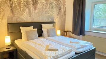 Un letto matrimoniale con lenzuola bianche, una testiera decorata con un motivo a foglia, un comodino con una lampada e una finestra con tende.
