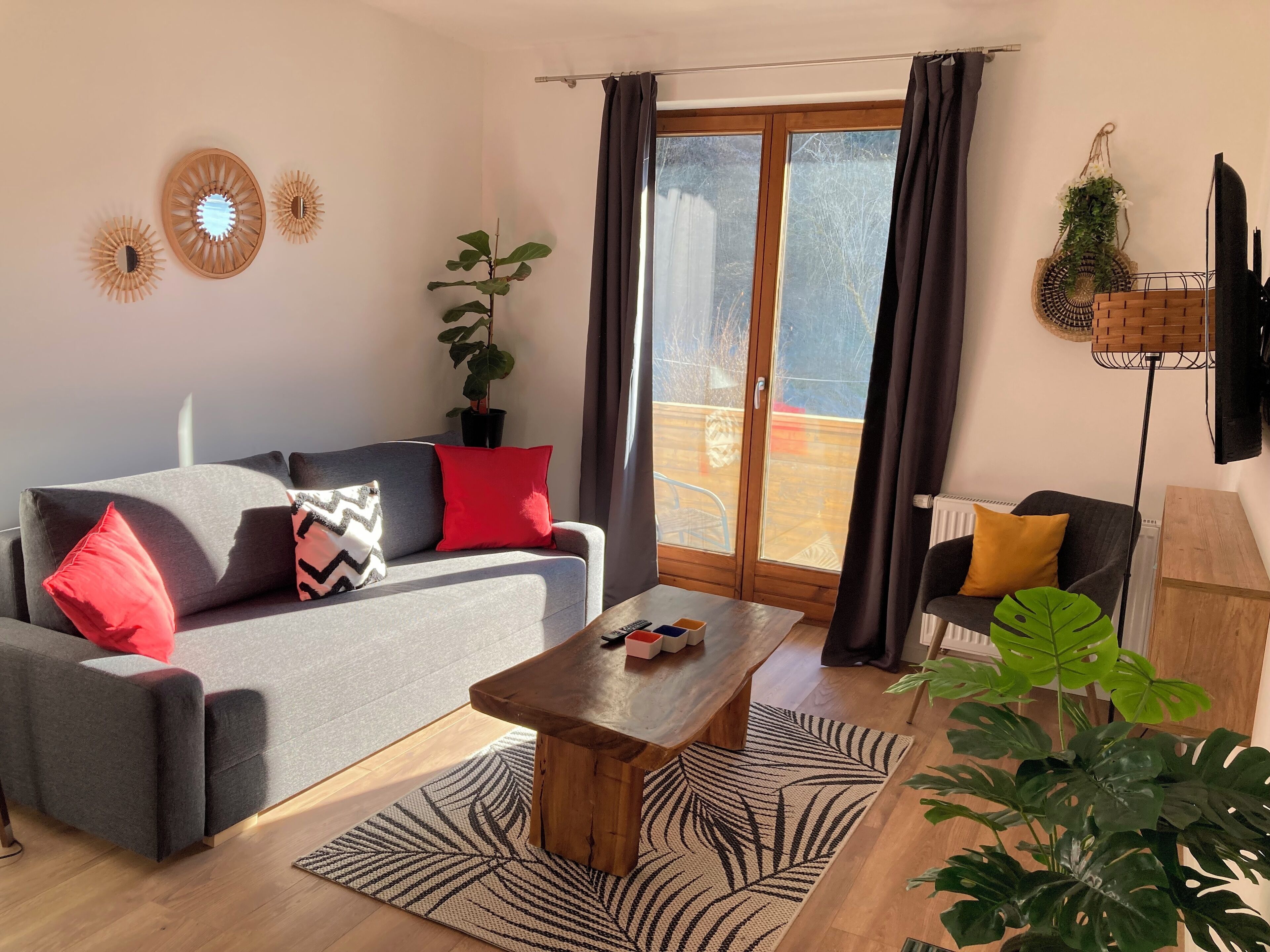 Un salon moderne avec un canapé gris, des coussins rouges et noirs, une table basse en bois et une grande fenêtre avec des rideaux.