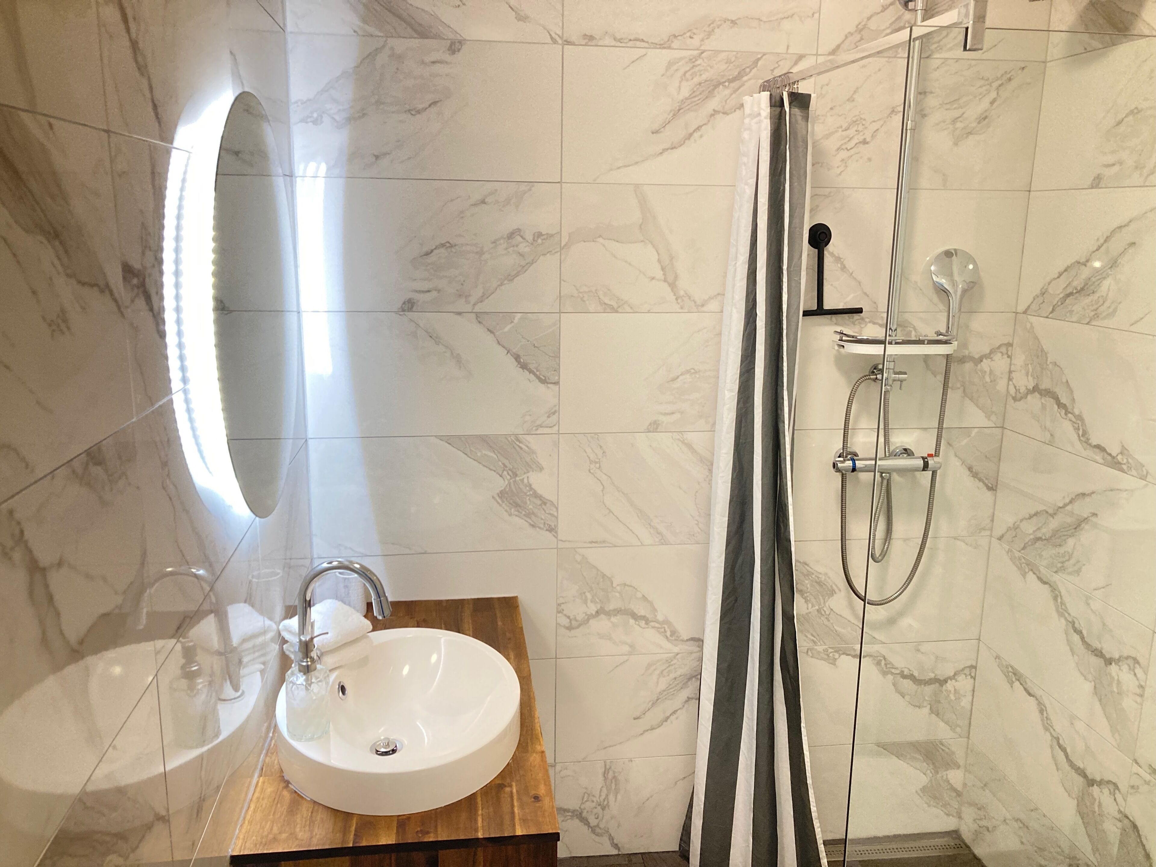 A bathroom with a marble-tiled wall, a wooden vanity with a sink, a mirror, and a shower with a handheld showerhead.