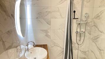 A bathroom with a marble-tiled wall, a wooden vanity with a sink, a mirror, and a shower with a handheld showerhead.