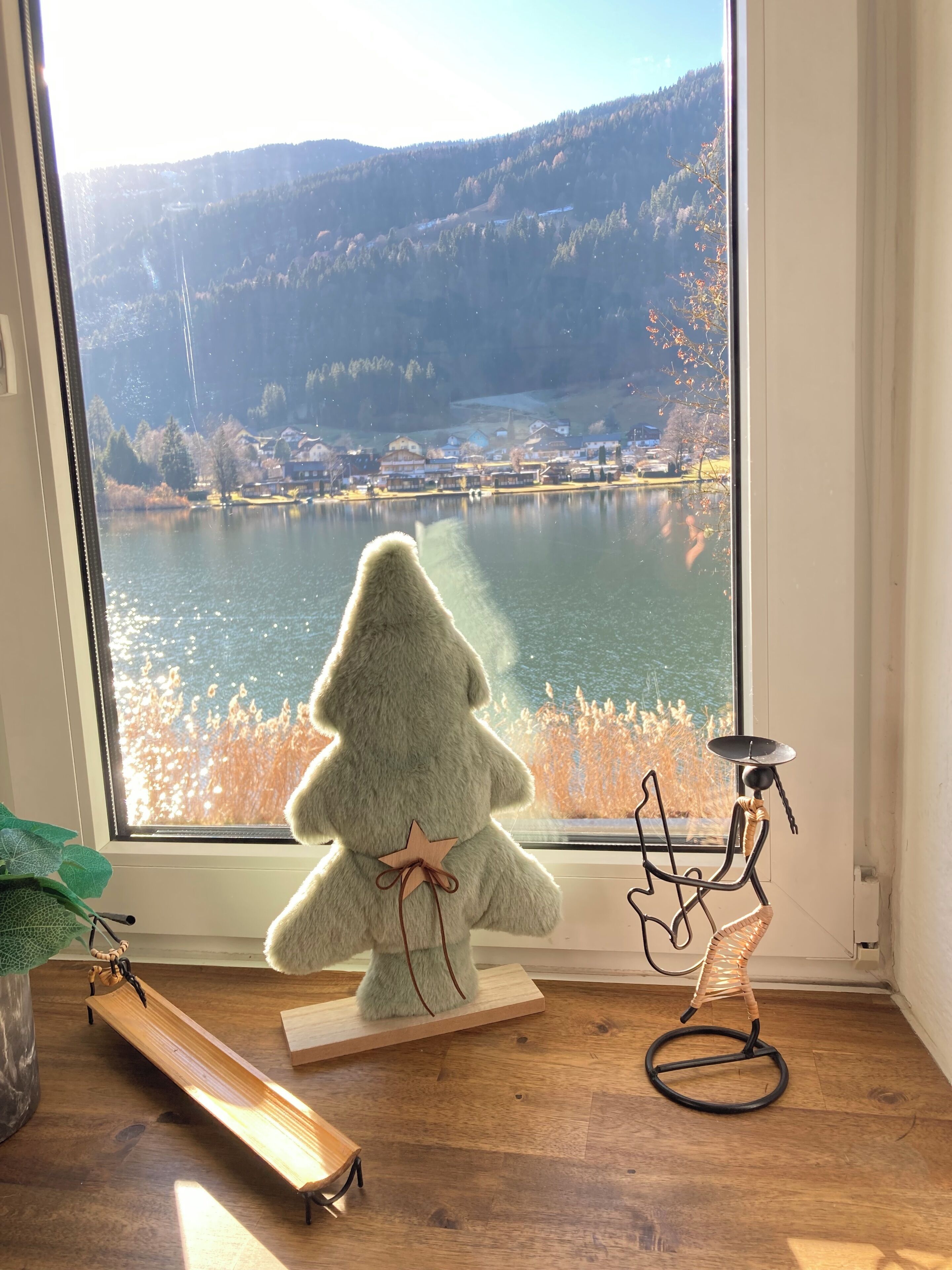 A plush Christmas tree on a windowsill with a view of a lake and mountains.
