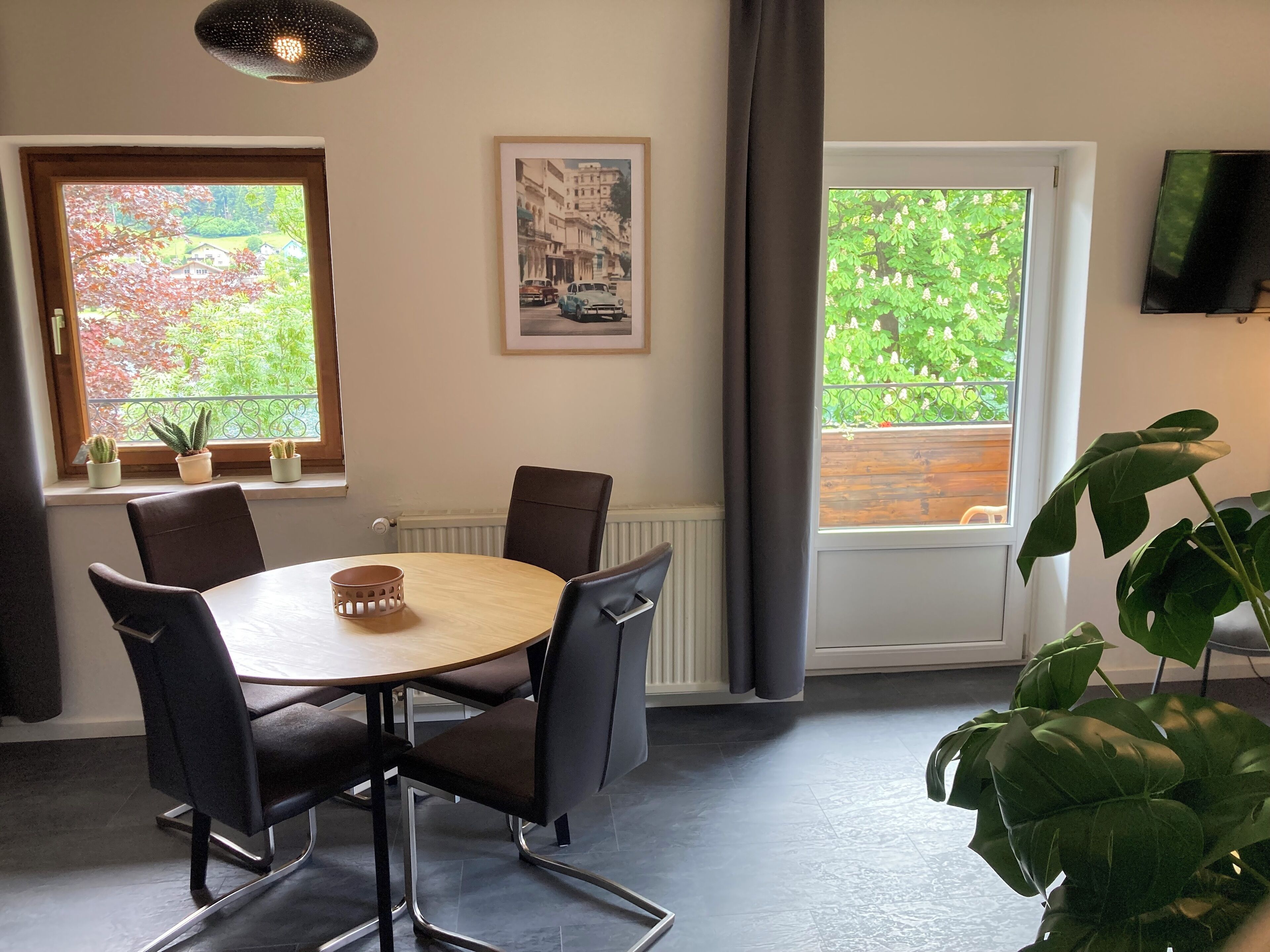 A dining area with a round table, four chairs, a potted plant, and two windows with views of greenery.