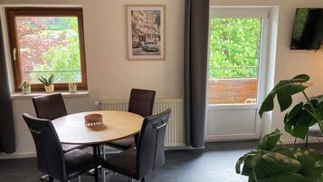 A dining area with a round table, four chairs, a potted plant, and two windows with views of greenery.