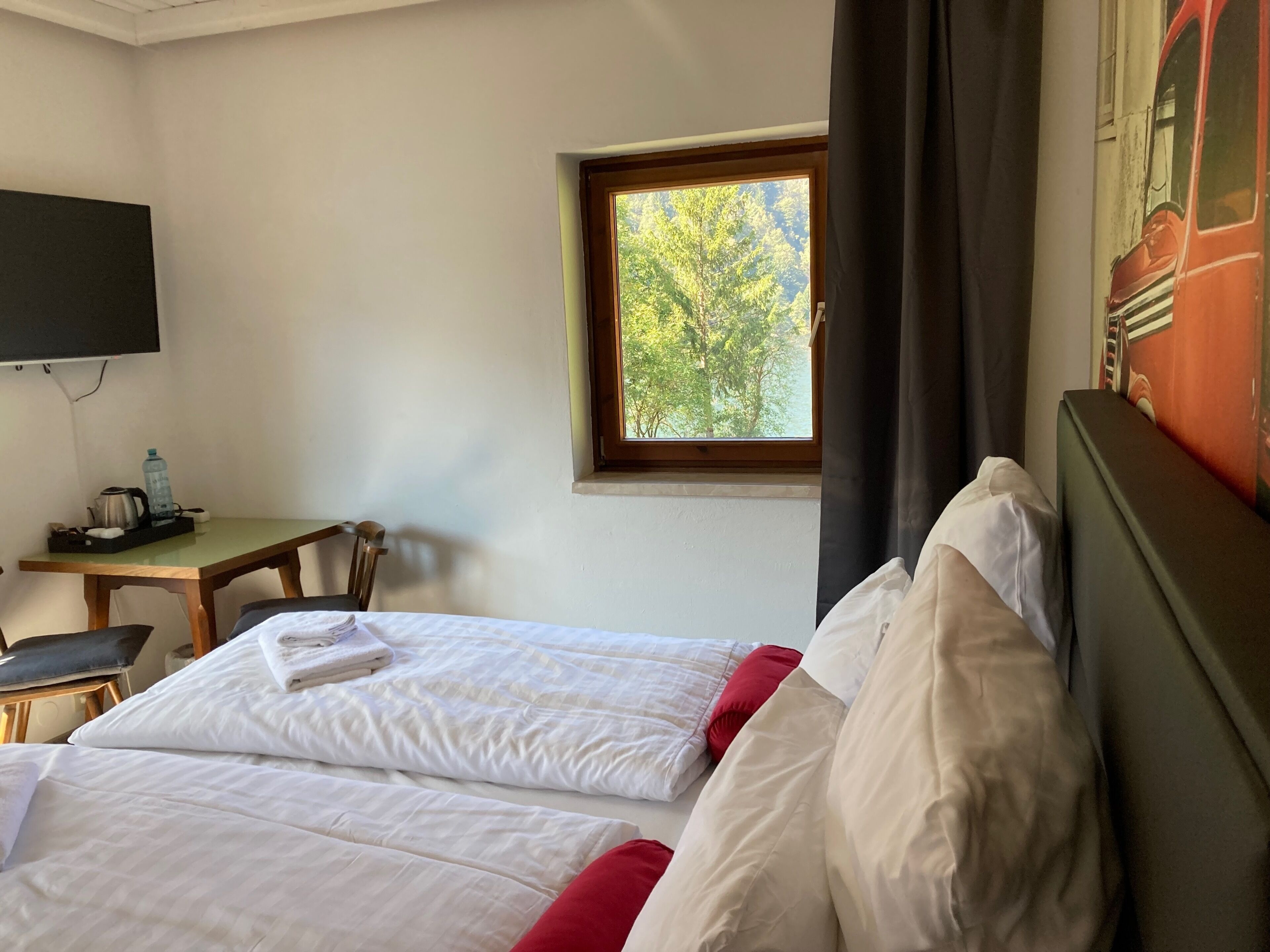 Ein Hotelzimmer mit zwei Betten, einem Schreibtisch mit Stuhl, einem an der Wand befestigten Fernseher und einem Fenster mit Blick auf Bäume.