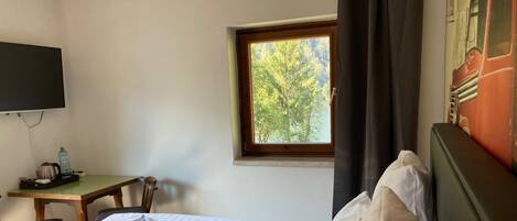 Ein Hotelzimmer mit zwei Betten, einem Schreibtisch mit Stuhl, einem an der Wand befestigten Fernseher und einem Fenster mit Blick auf Bäume.