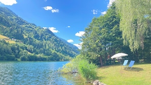 Vista al lago con agua cristalina, un área verde con dos sillas azules y una sombrilla blanca, y un promontorio boscoso al fondo.