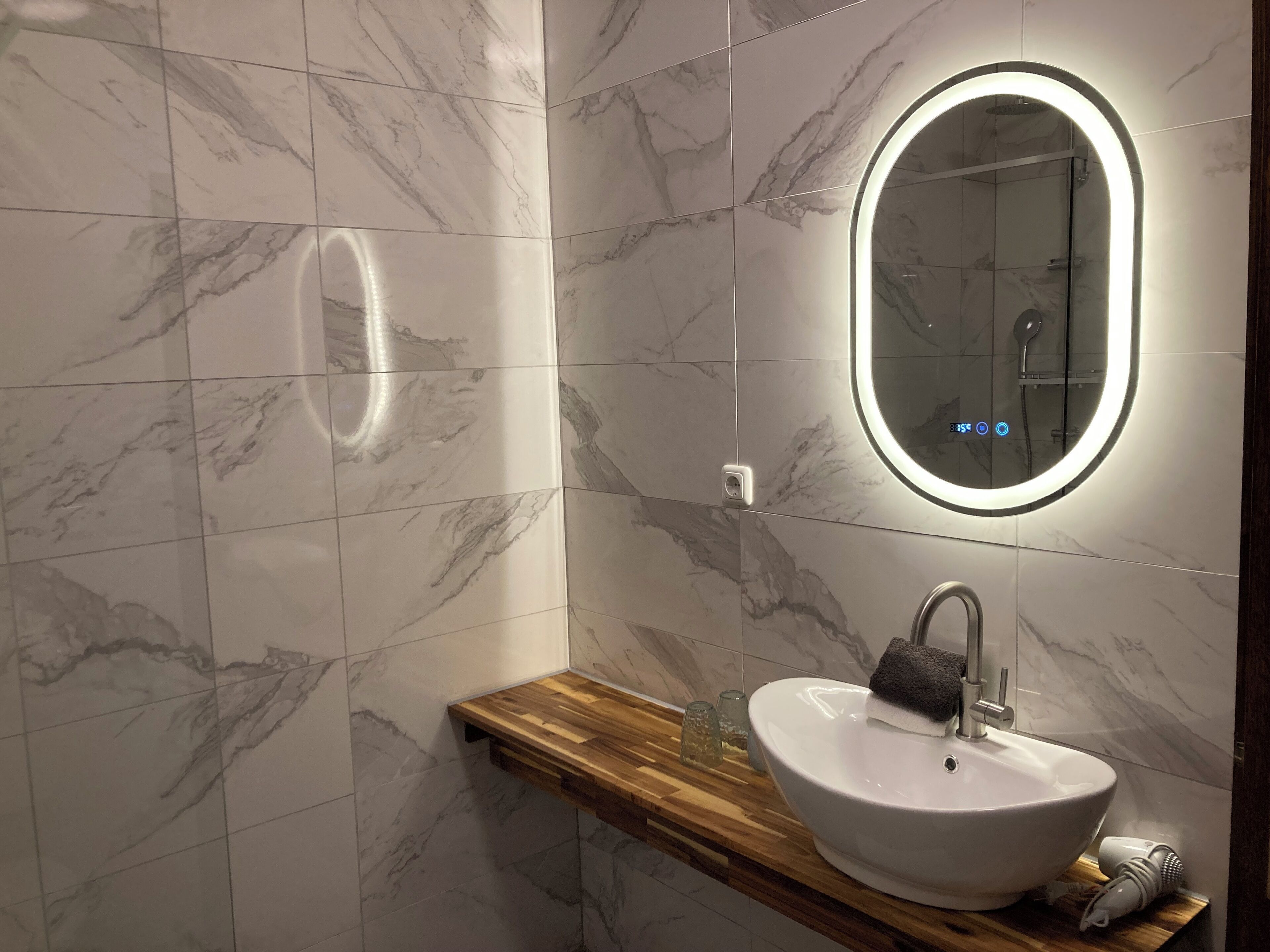 A modern bathroom with marble tiles, a wooden vanity, a white sink, a mirror with a light, and a shower.