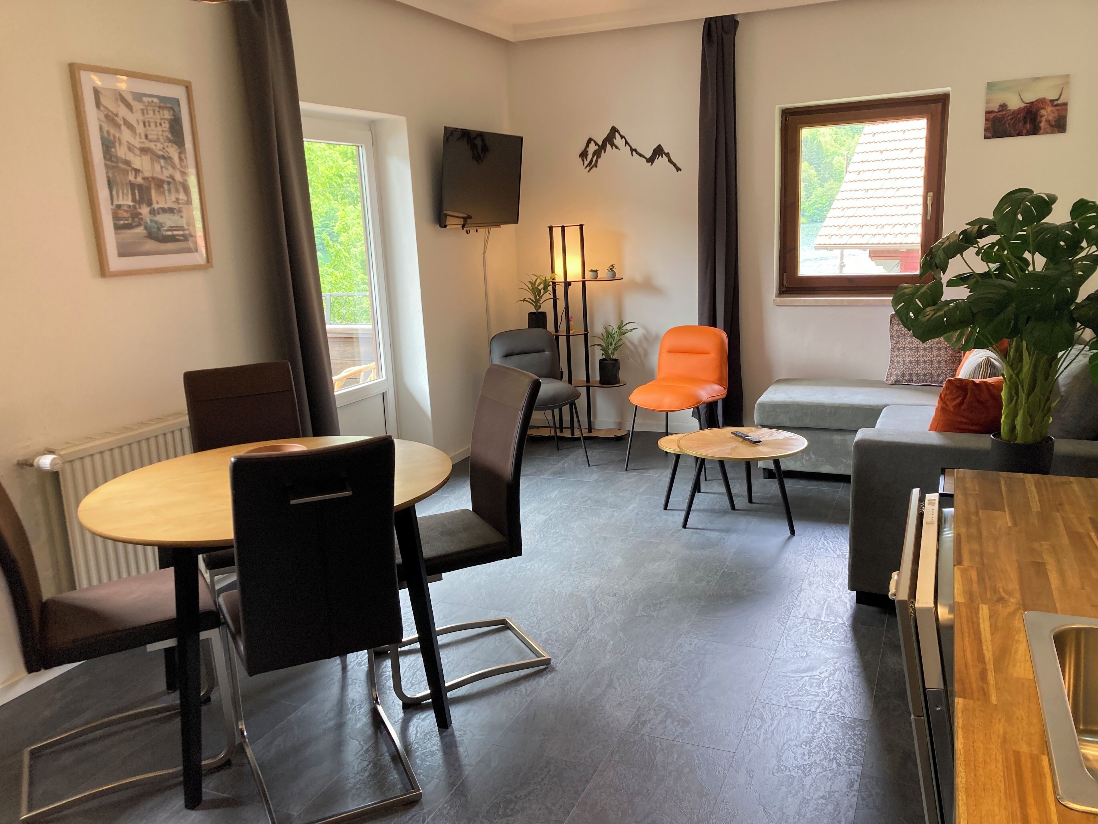 A modern living room with a dining area, a grey sofa, an orange chair, a wooden table, and a TV mounted on the wall.