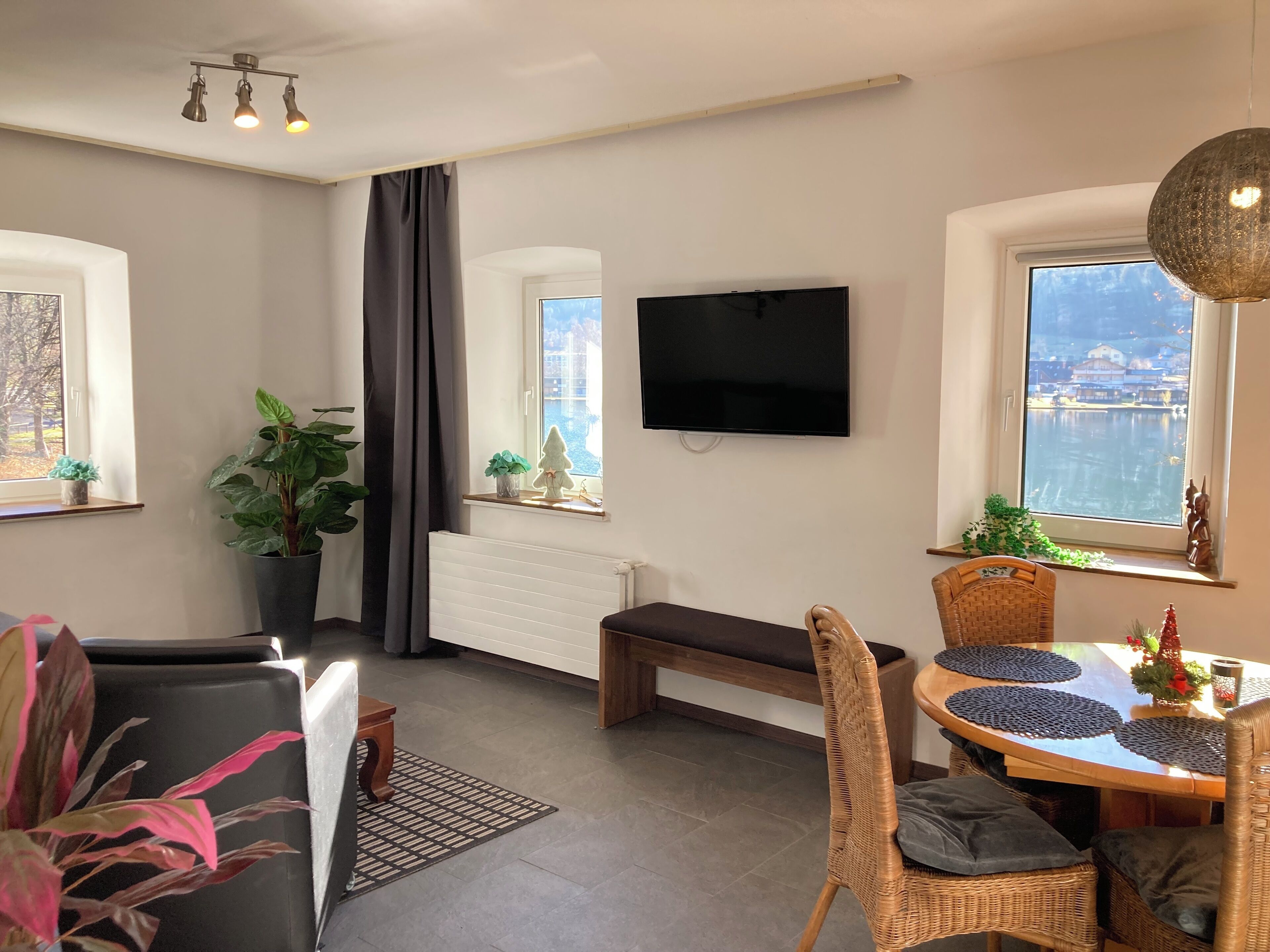 A modern living room with a large window, a flat-screen TV, a dining area with wicker chairs, and a potted plant.