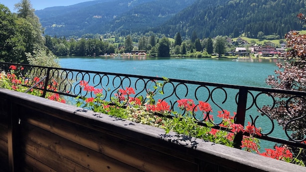 Una vista panorámica de un lago con montañas al fondo, una terraza de madera con barandilla de metal y flores rojas en primer plano.