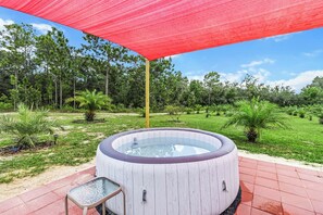 Un bain à remous avec un revêtement extérieur en bois, entouré d’un auvent rouge, installé sur une terrasse avec une table et des chaises en verre, et une zone herbeuse avec des arbres en arrière-plan.
