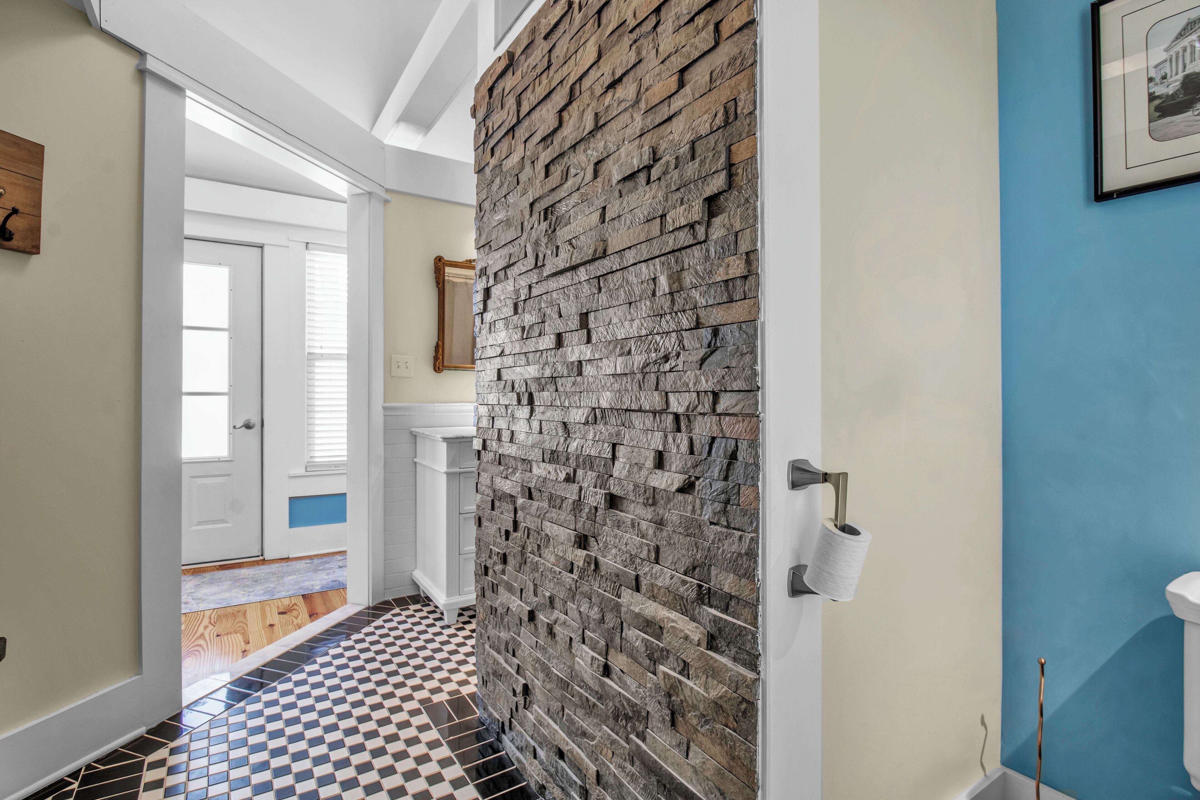 A hallway with a checkered floor, a stone wall, a white door, and a blue wall with a framed picture.