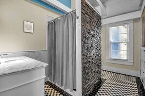 Un baño con pared de piedra, suelo a cuadros y ventana con persianas.