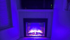 A room with a fireplace, a television mounted above it, and a window with blinds. - Lakefront home away from home! (Hot Springs)