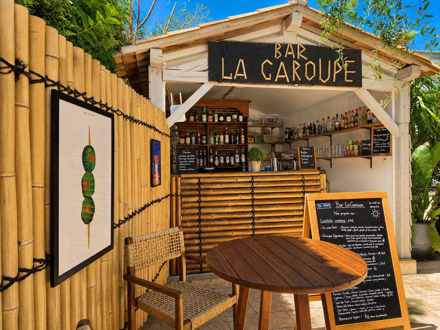 A small outdoor bar named 'Bar La Garoupe' with a bamboo fence, a wooden table, a wicker chair, and various bottles and glasses on shelves.