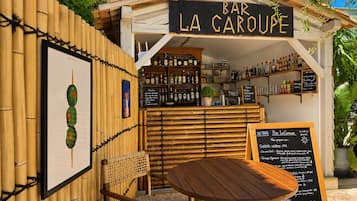 A small outdoor bar named 'Bar La Garoupe' with a bamboo fence, a wooden table, a wicker chair, and various bottles and glasses on shelves.