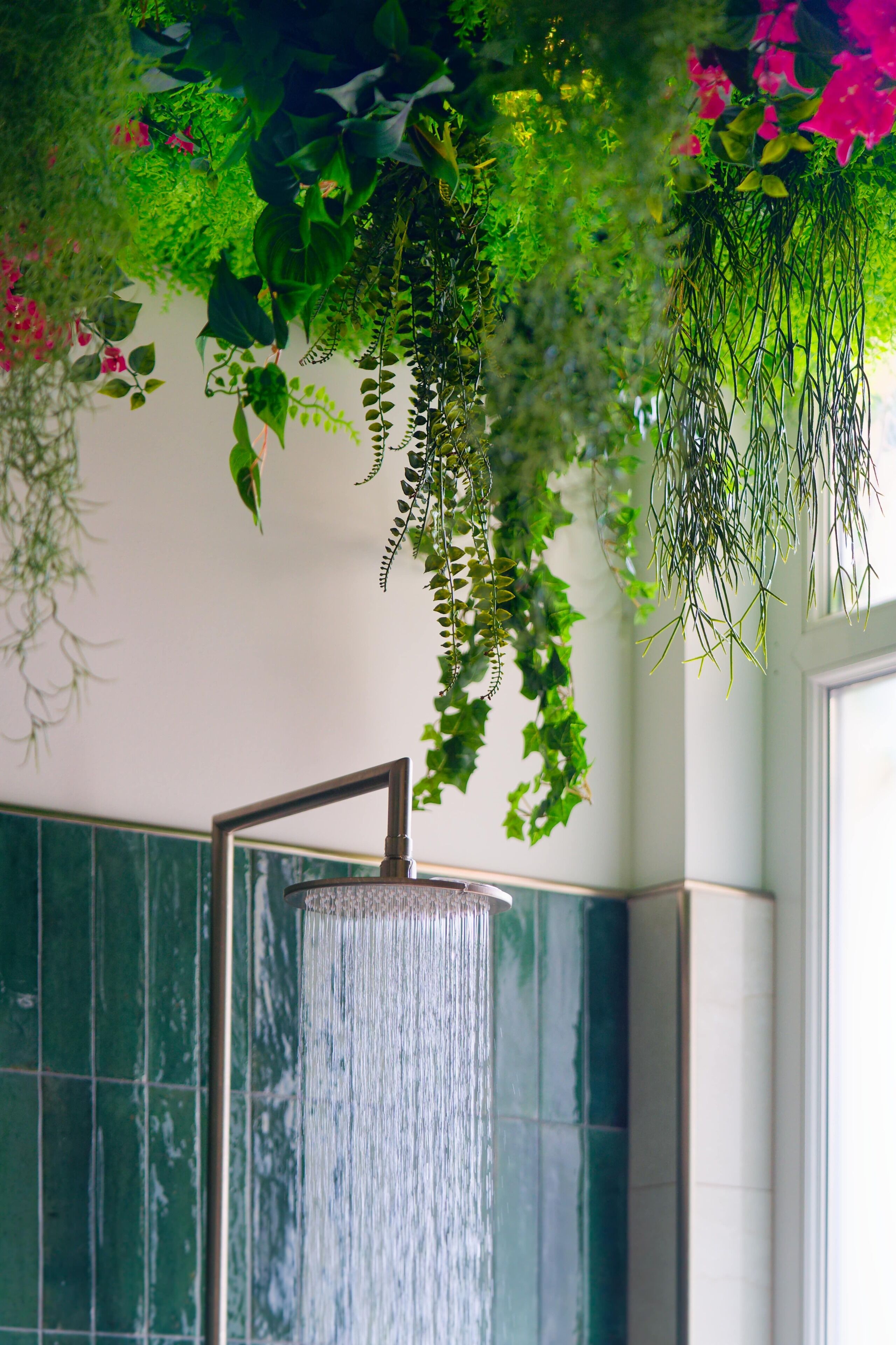 A shower with a vertical water flow and green plants hanging above.