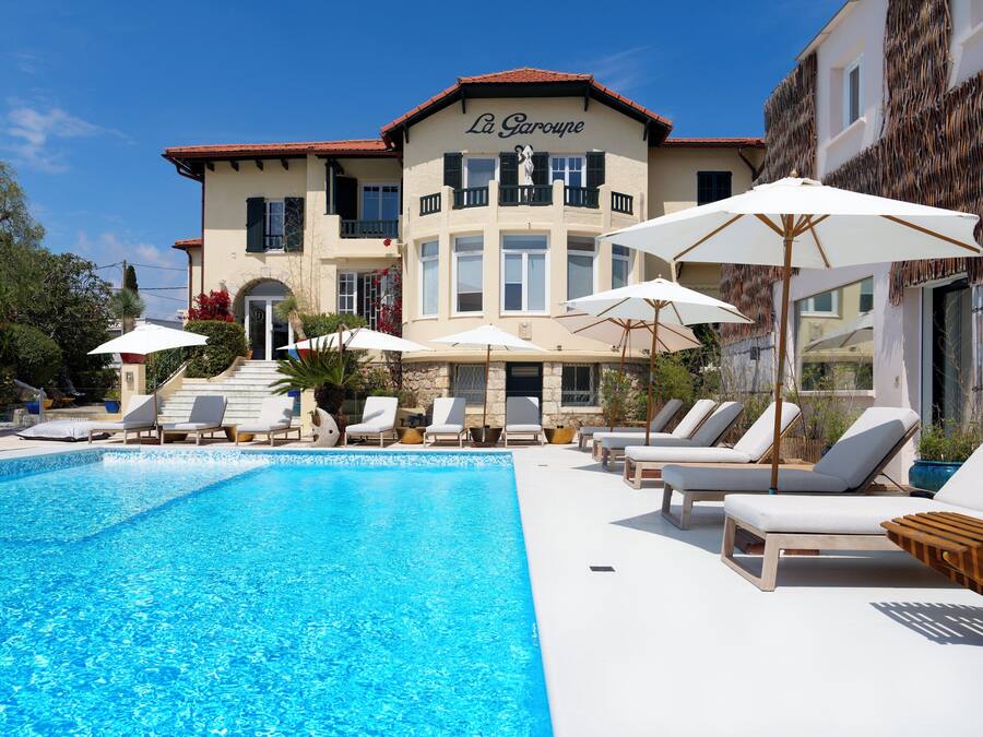Una piscina con sillas de descanso y sombrillas, un edificio con un letrero que dice "La Garoupe", y un cielo azul despejado.