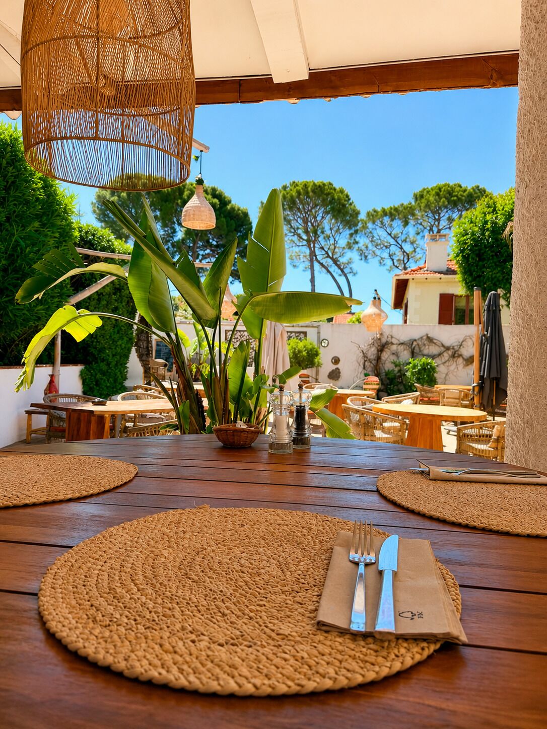 Un espace de restauration en plein air agrémenté de sets de table en fibres tressées, d’un luminaire suspendu en osier et offrant une vue sur un bâtiment doté d’un toit rouge.