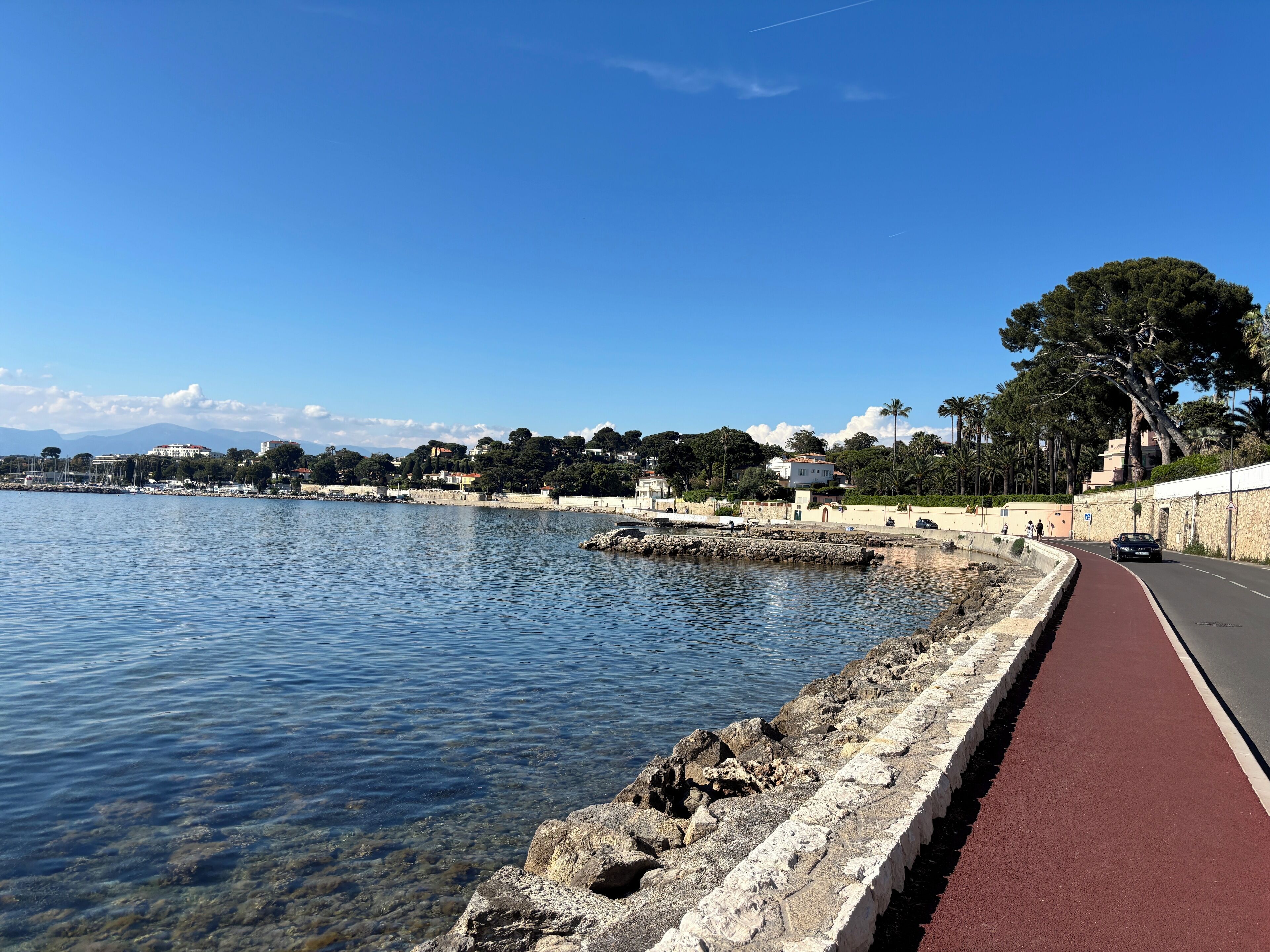 Une route côtière avec une piste cyclable rouge, une eau d’un bleu limpide et des bâtiments qui bordent la côte.