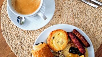Un plato de desayuno con bollería, salchichas y fruta, una taza de café y cubiertos sobre un posavasos tejido.