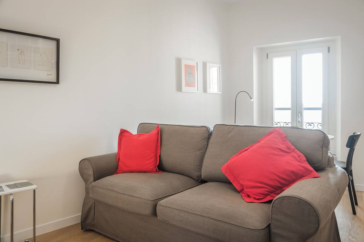 Een moderne woonkamer met een bruine bank, twee rode kussens, een klein wit bijzettafeltje en kunstwerken in lijsten aan de muur.