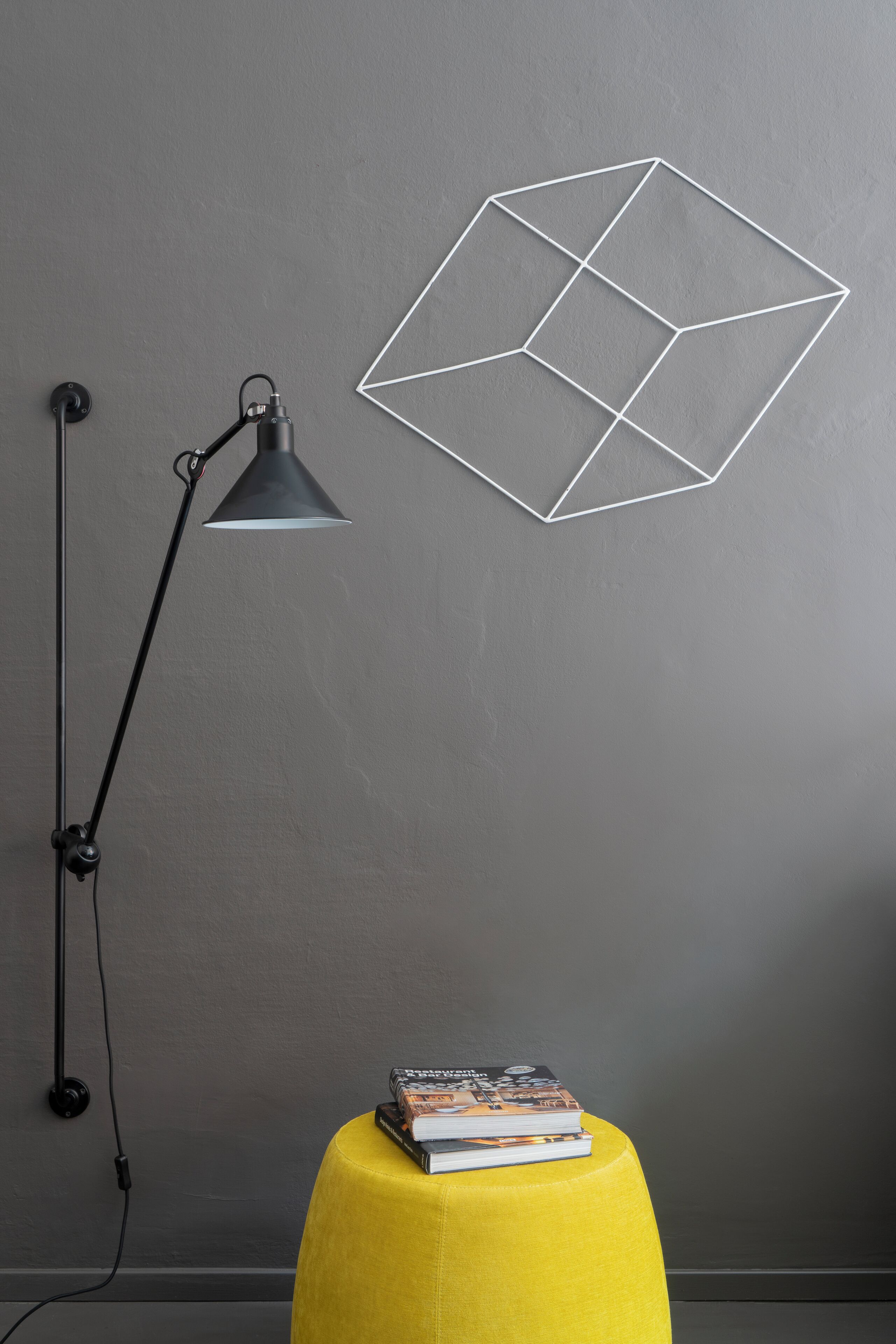 A room with a geometric wall art piece, a black desk lamp, and a yellow stool with books.
