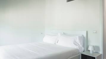 Un dormitorio minimalista con una cama individual, ropa de cama blanca, una mesita de noche con lámpara y una pared lisa.
