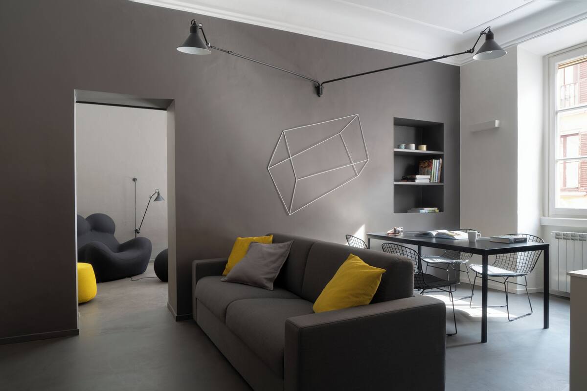Een moderne woonkamer met een grijze bank, gele kussens, een eettafel met stoelen en een geometrische lamp aan de muur.