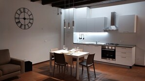Une cuisine moderne avec des armoires blanches, un four encastré et des ustensiles de cuisine suspendus. On y trouve une table à manger pour quatre personnes, avec des chaises. Une grande horloge murale est visible.