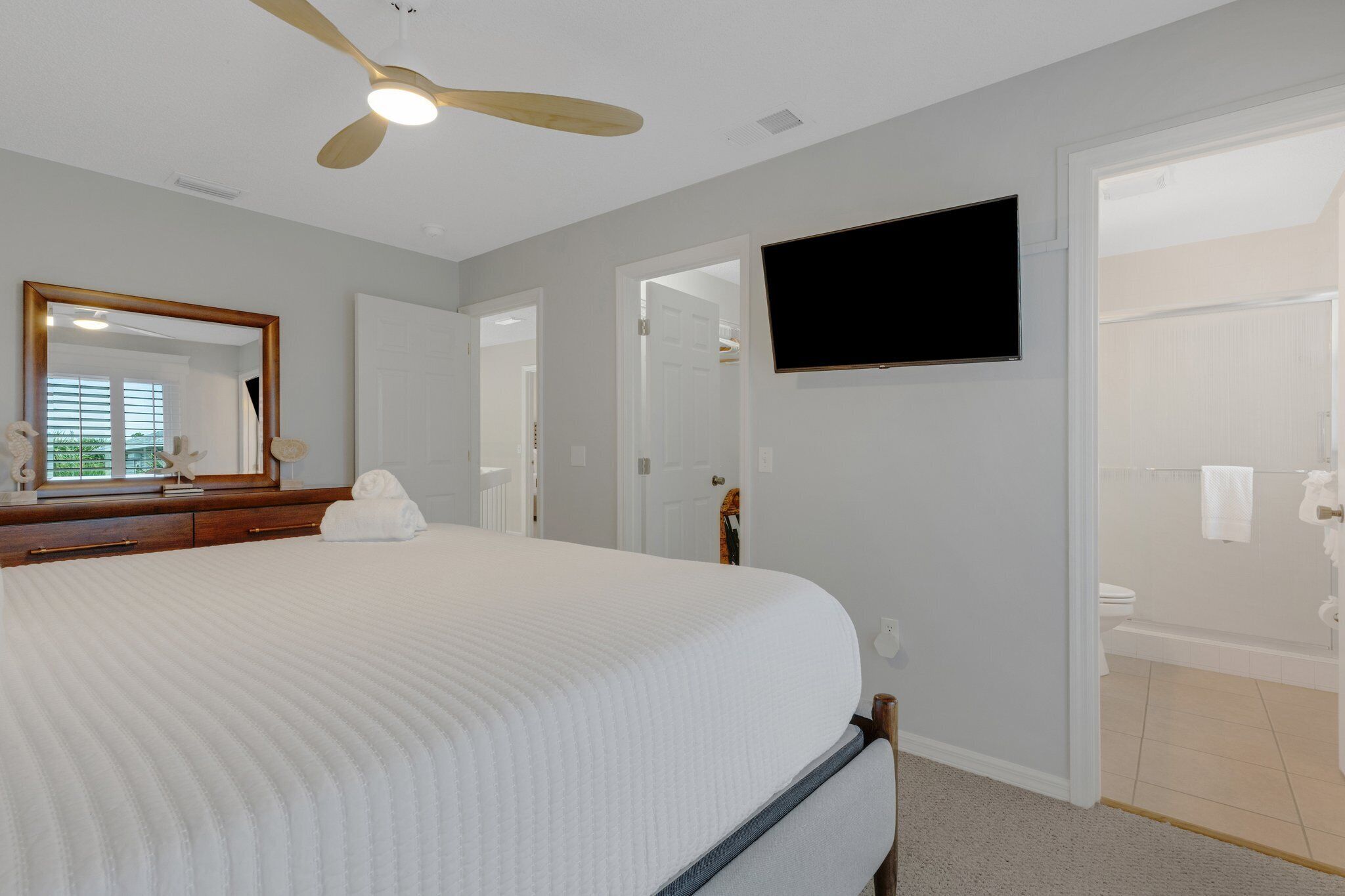 A bedroom with a large bed, a ceiling fan, a TV mounted on the wall, and a bathroom visible through an open door.