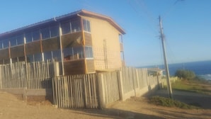 Edificio de dos pisos con un balcón en el nivel superior, cerca hay una valla de madera y se ve el océano con claridad.