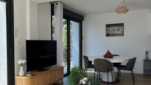 A modern living room with a flat-screen TV, wooden entertainment unit, glass coffee table, and a dining area with a white table and chairs.